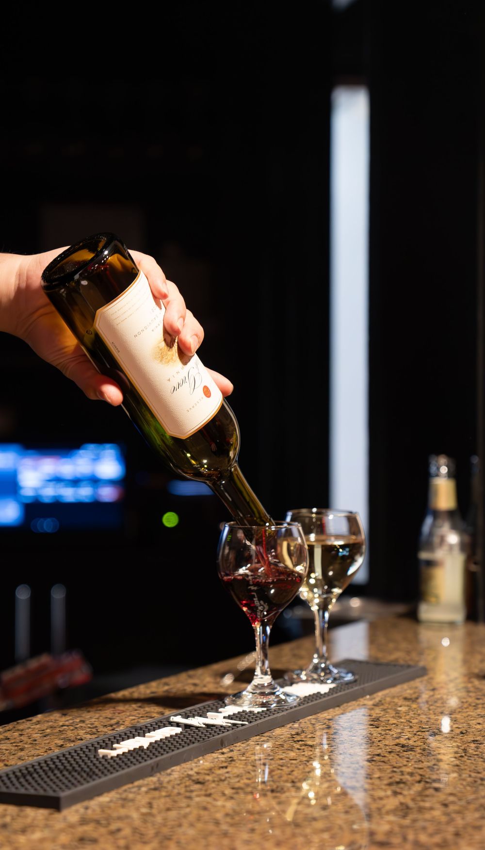 Bartender pouring red wine from a bottle into a glass on a granite bar counter in a dimly lit venue.