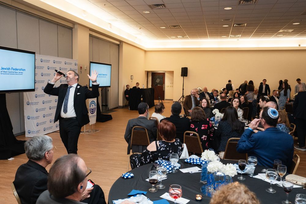 Speaker gestures at Jewish Federation gala event with guests seated at decorated tables in a banquet hall.