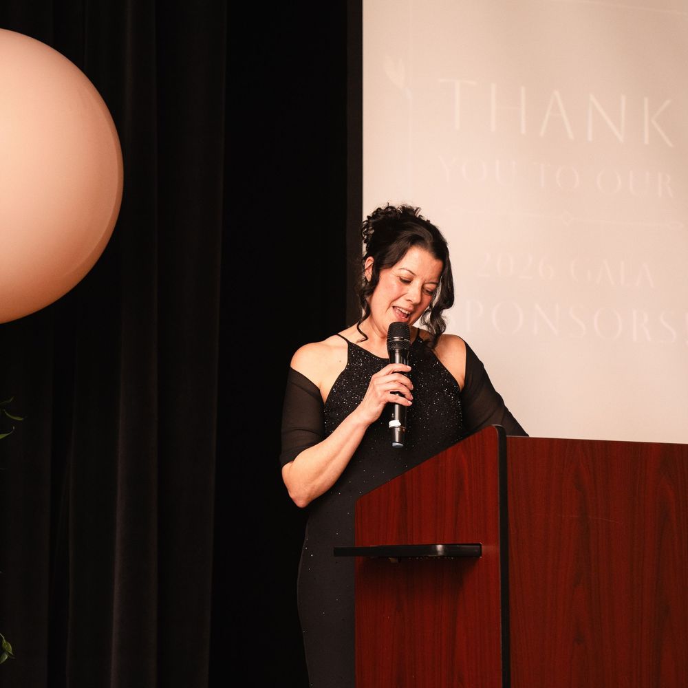 Woman in black dress speaks at podium with microphone at formal thank you donor recognition event.