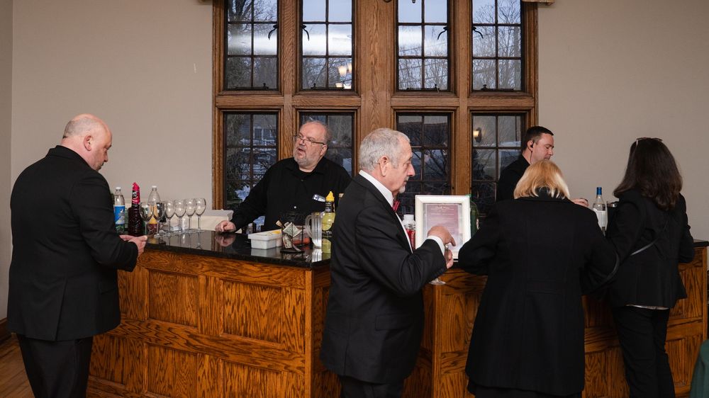 Guests in formal attire gather around a wooden bar during an indoor event, with bottles and glassware displayed.