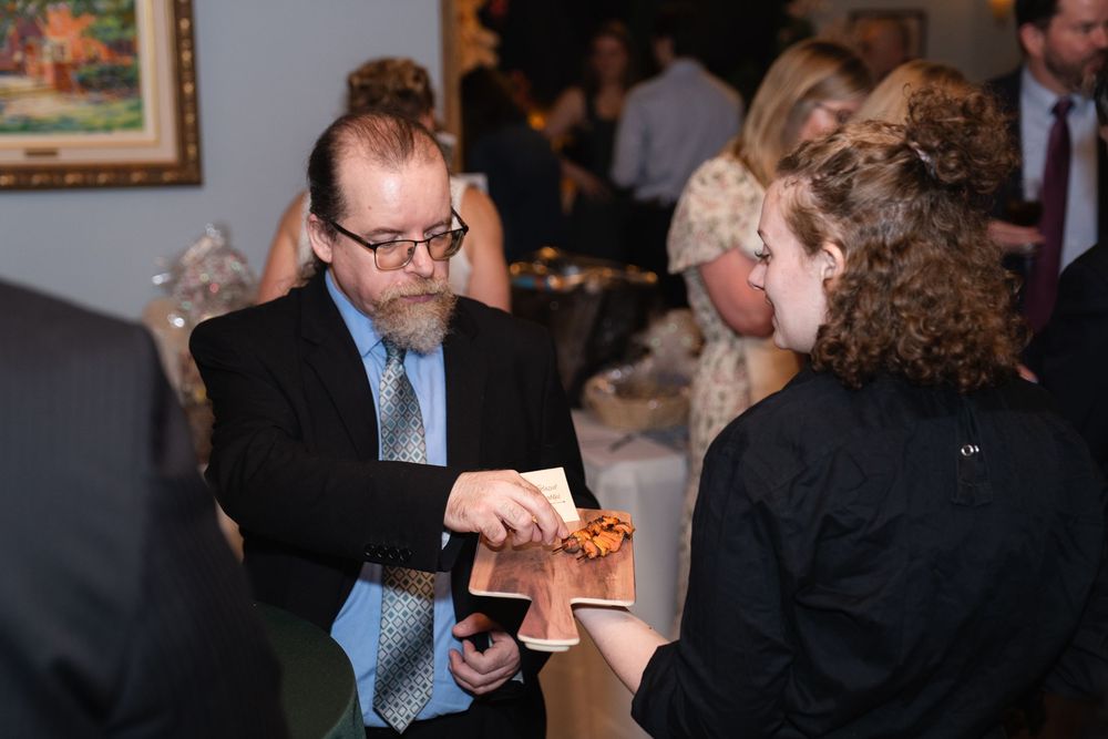 Man in suit and tie serves food at an elegant indoor catered event with guests mingling in background.