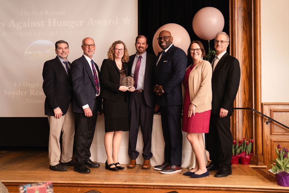 Group of seven professionals posing on stage at a Fight Against Hunger Award ceremony with balloon decor.