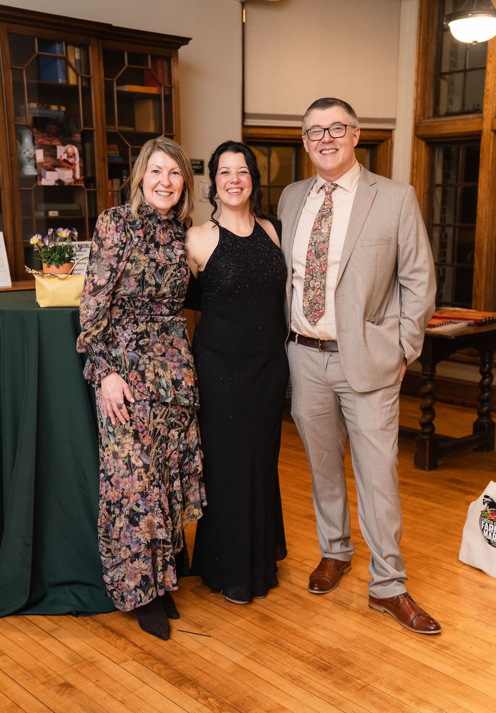 Three elegantly dressed people smiling together at a formal event in a warmly lit room with hardwood floors.