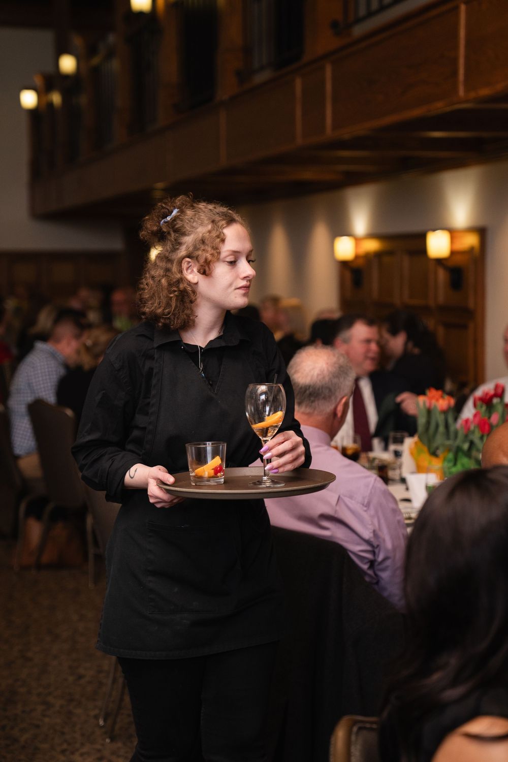 A waitress in black uniform carries a tray with drinks through a busy upscale restaurant dining room.
