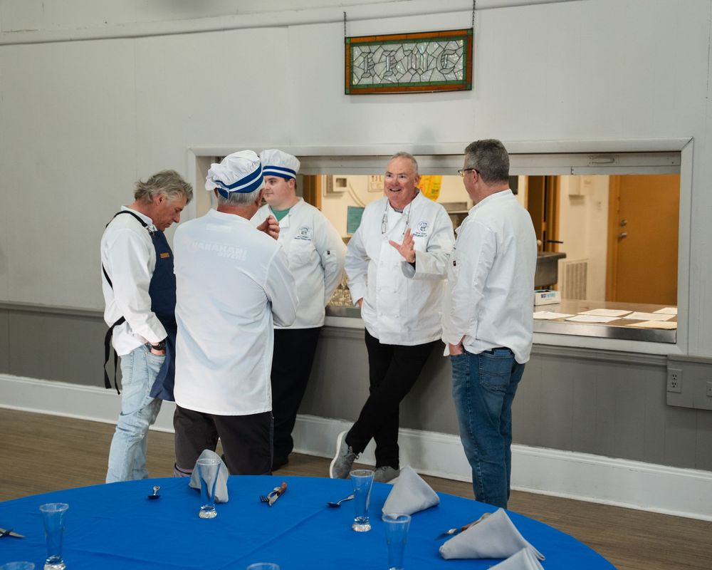 Group of chefs in white uniforms discussing around a blue table set for an event in a hall.