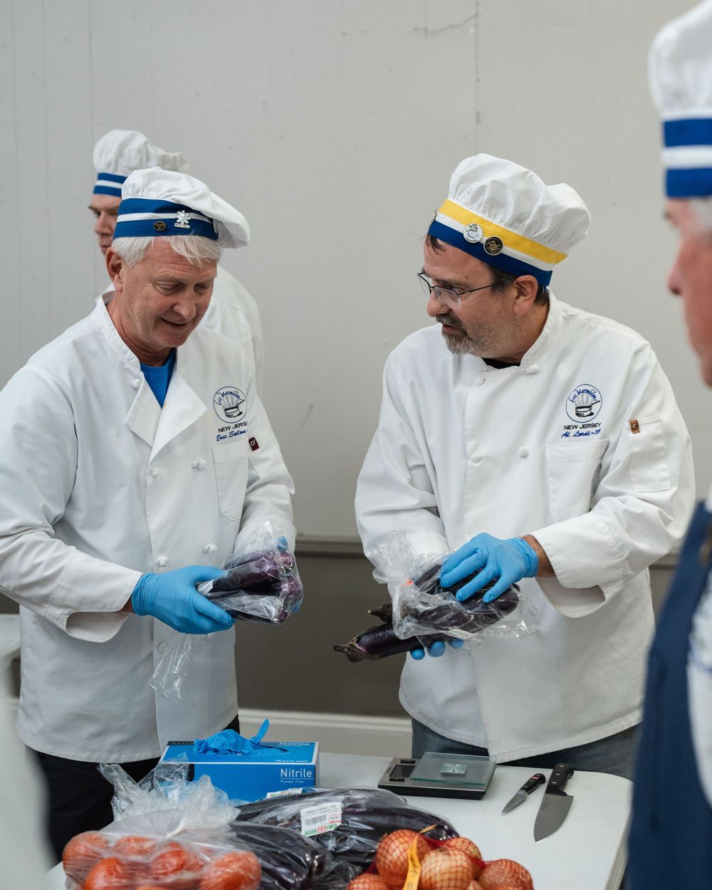 Two chefs in white uniforms and hats handle packaged food items at a preparation table with fresh produce.