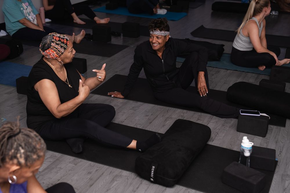 Two women in black activewear laugh together during a group yoga class on dark mats in a modern studio.