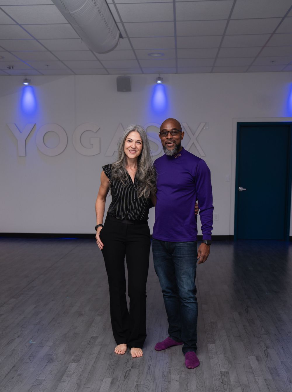 Two smiling people pose together inside a YogaFX studio with blue accent lighting and hardwood floors.