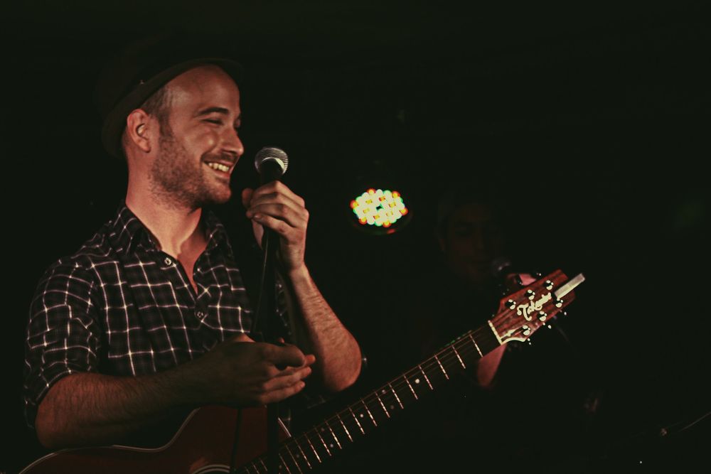 Smiling male musician performing live on stage holding a microphone and acoustic guitar under warm stage lighting.