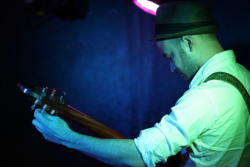 Male guitarist in a fedora and suspenders playing acoustic guitar under blue and green stage lights.