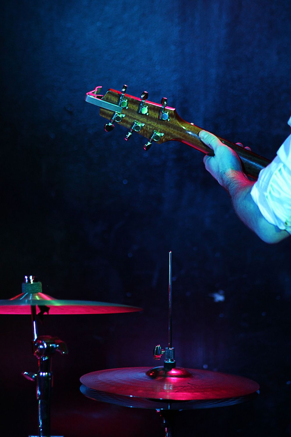 Close-up of a guitarist holding an electric guitar neck above a drum kit on a dark stage with colorful lighting.