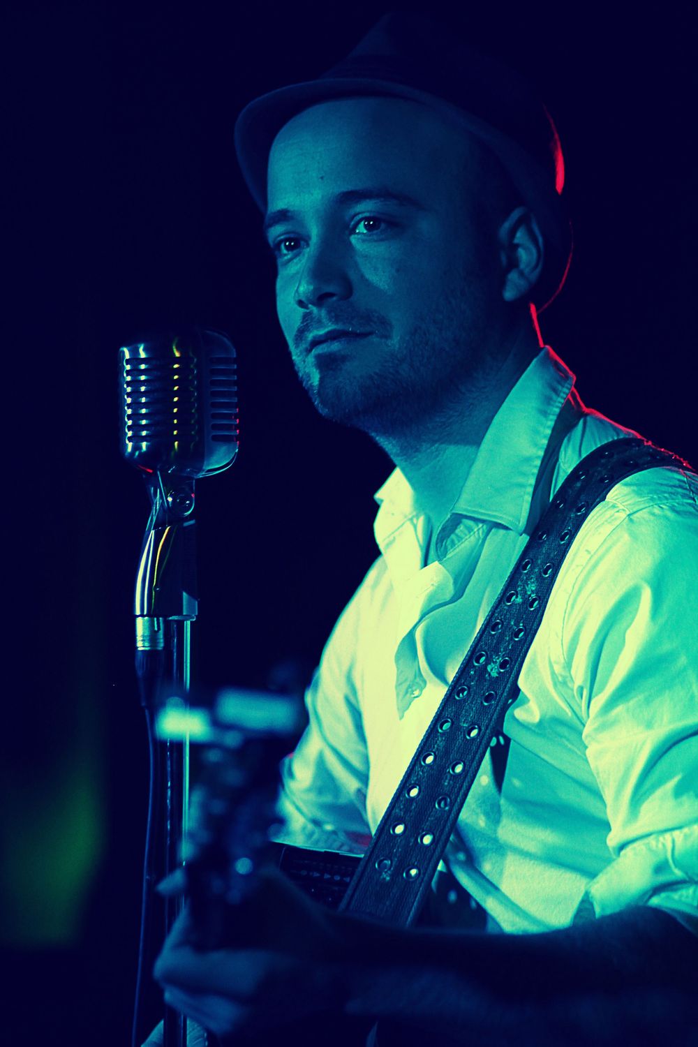 Male musician in a white shirt and hat performing with a guitar near a vintage microphone under blue and green stage lights.
