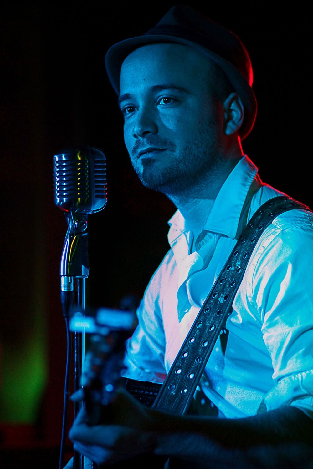 Male guitarist in a hat and white shirt stands by a retro microphone under dramatic blue and red stage lighting.