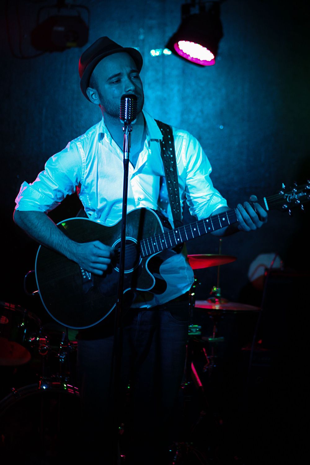 Male musician singing and playing acoustic guitar on stage under blue and red stage lights with drums visible behind him.