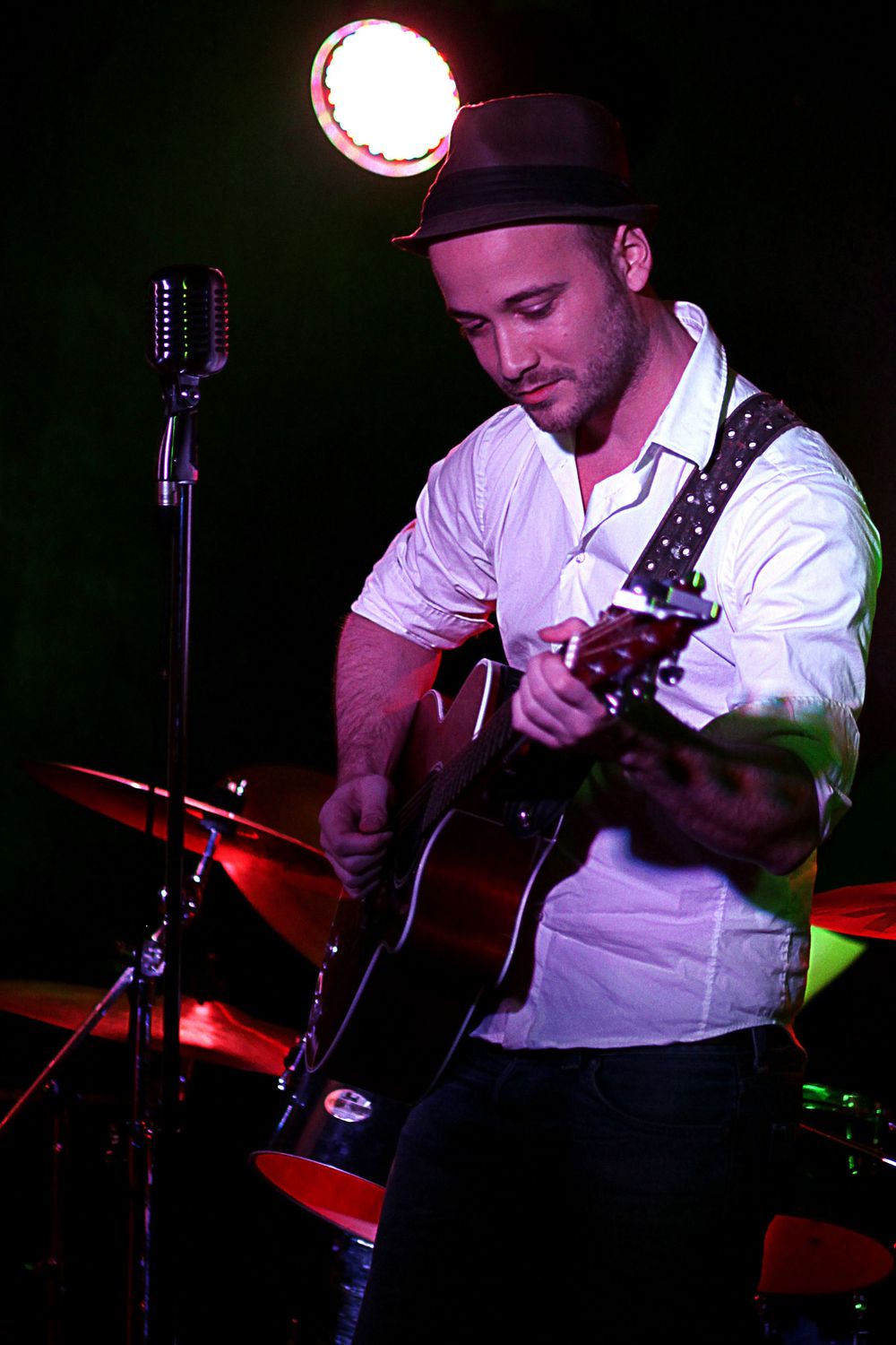 Male guitarist in a black hat and white shirt playing an acoustic guitar on stage under a bright spotlight.