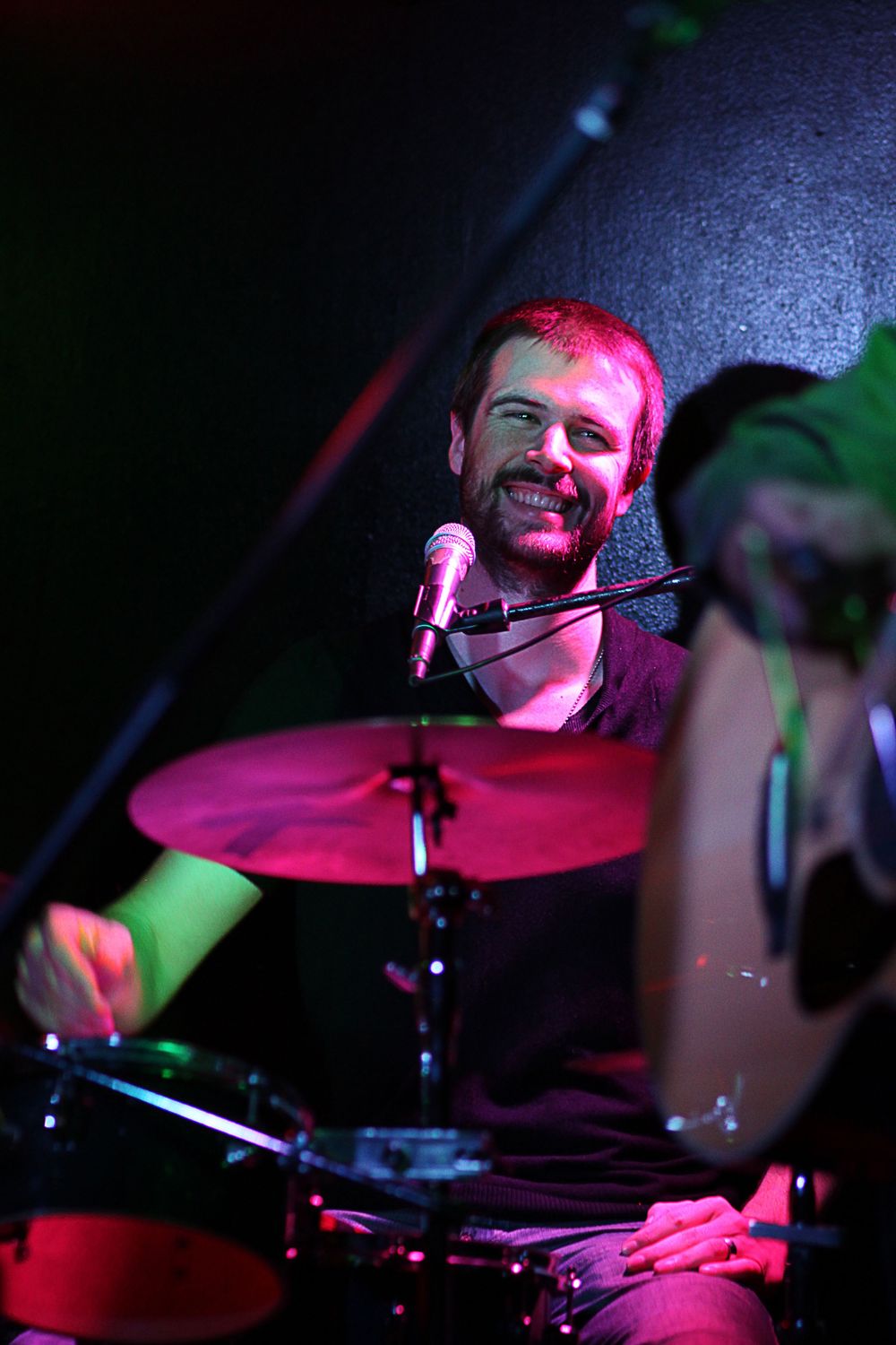 Smiling male drummer performing behind a drum kit under red stage lighting at a live concert.