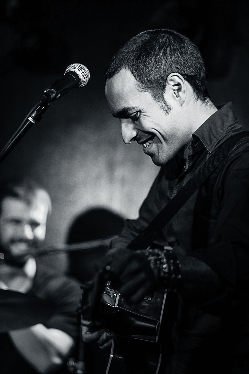 Black and white photo of a smiling male guitarist performing on stage with a microphone stand.