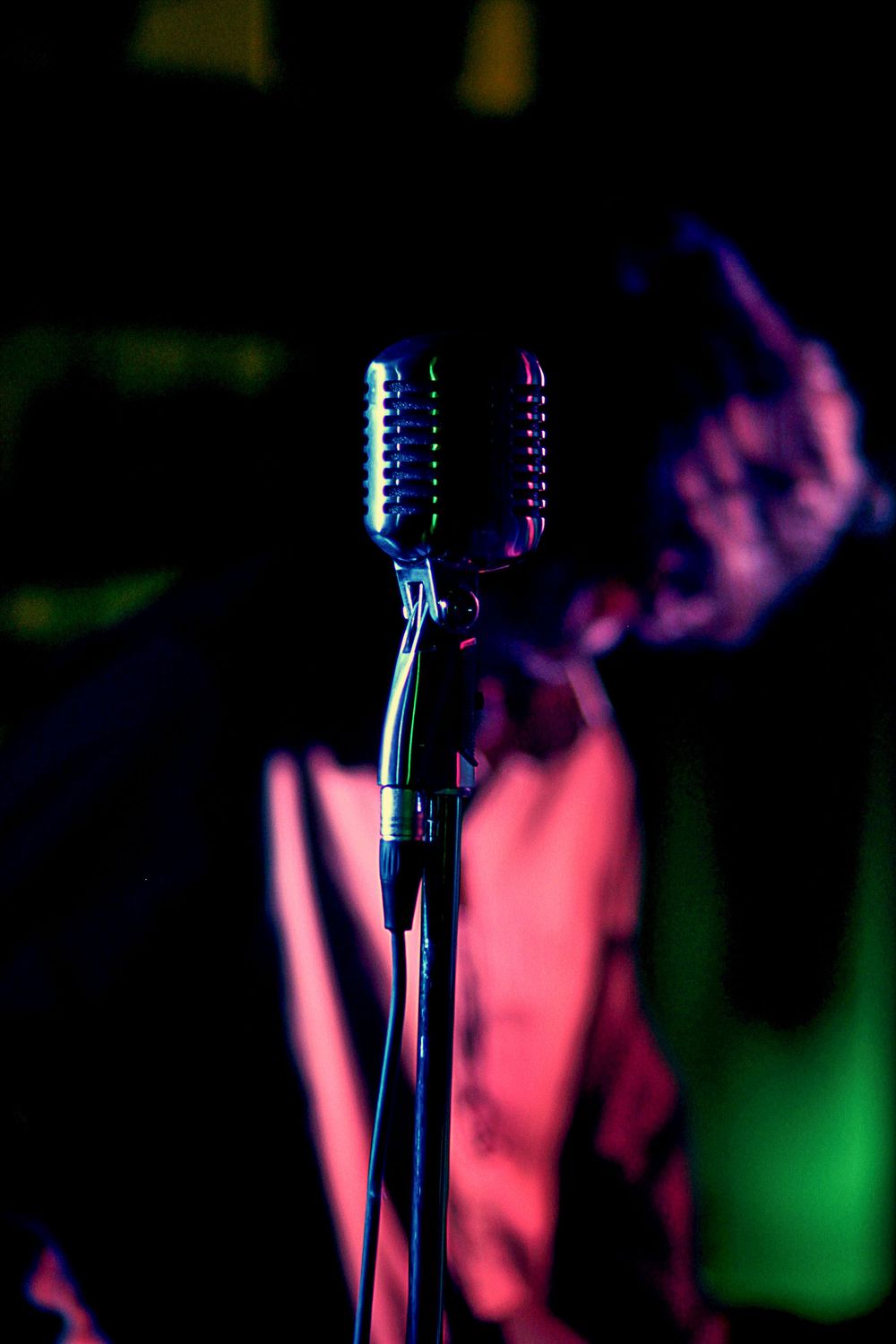 Vintage chrome microphone on a stand with colorful blue and pink stage lighting in the background.