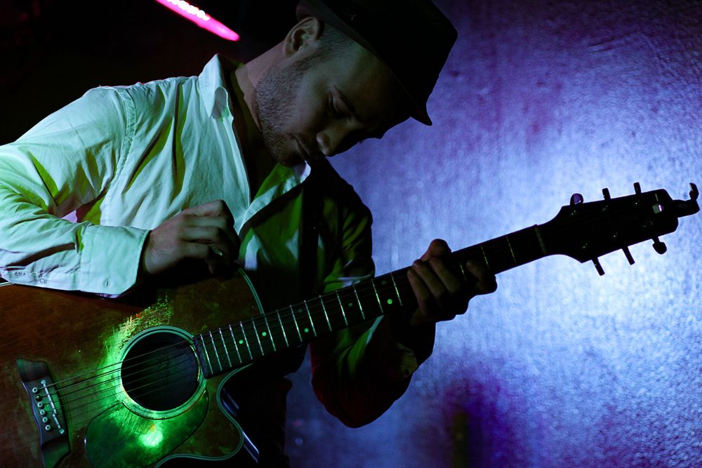 Male guitarist in a hat and white shirt playing acoustic guitar under green stage lighting.