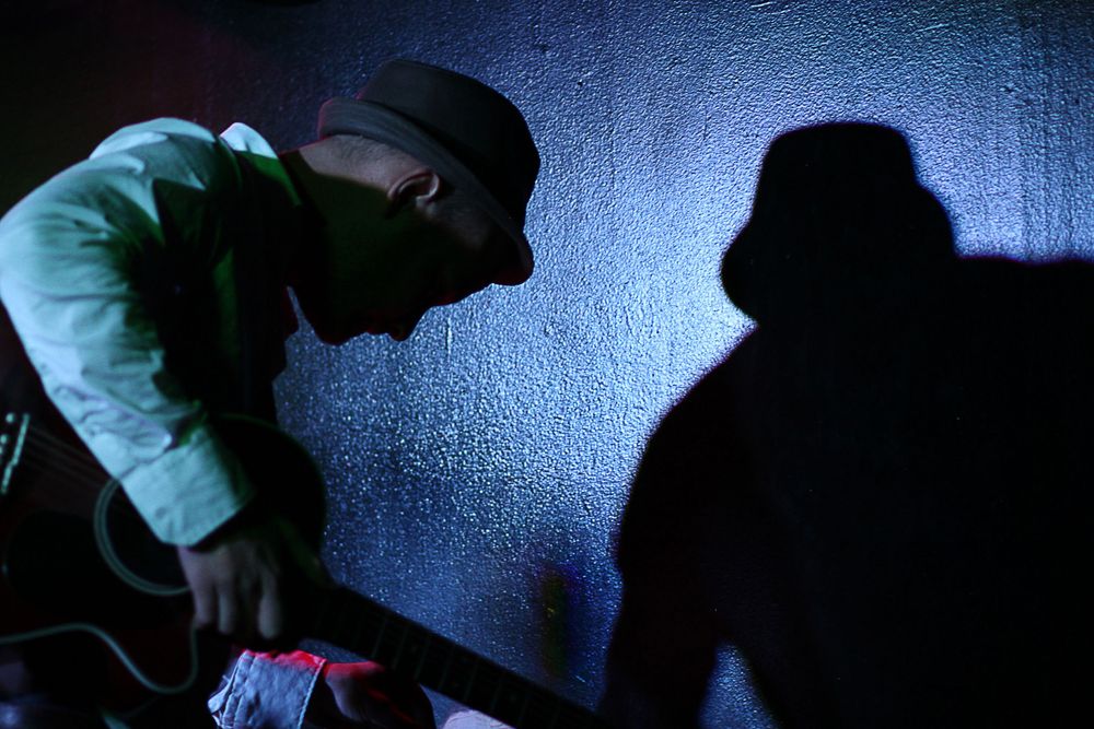 Male musician in a hat leaning over equipment with dramatic shadow cast on a blue-lit wall.