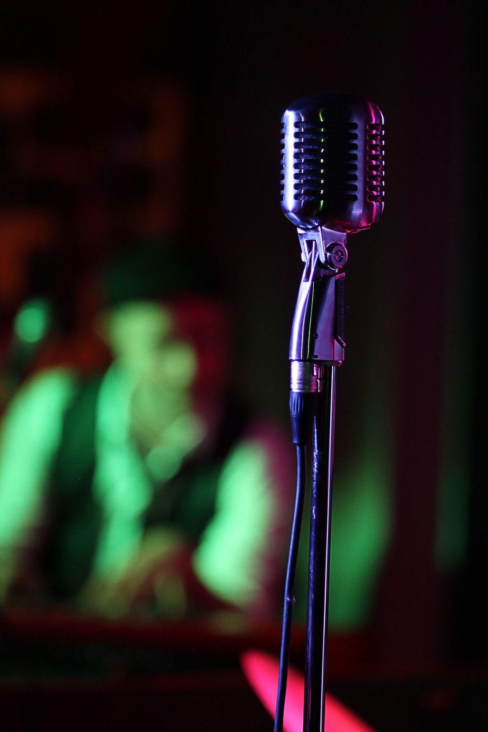Vintage silver microphone on a stand with a blurred guitarist visible in colorful stage lighting.