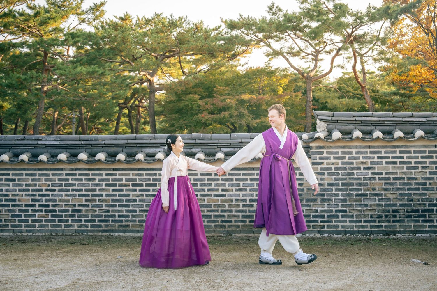 A scenic view of traditional Korean architecture with stone walls and autumn foliage in warm golden light.