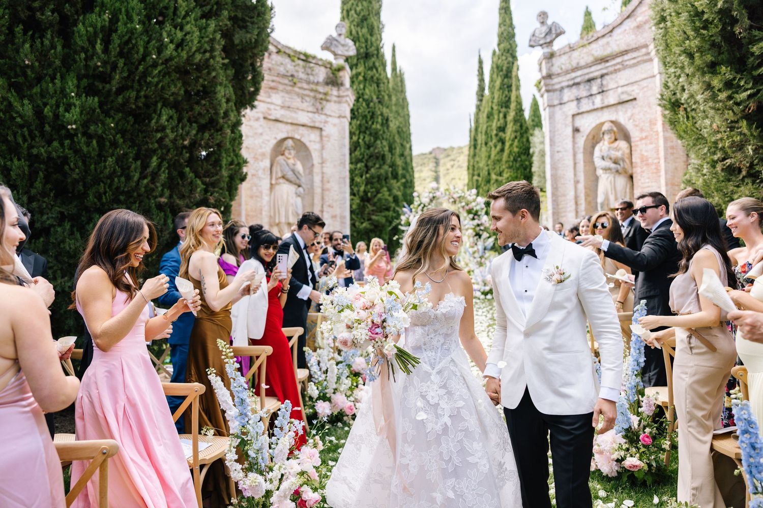 Wedding guests celebrate and dance together at an elegant outdoor reception with cypress trees and stone architecture.