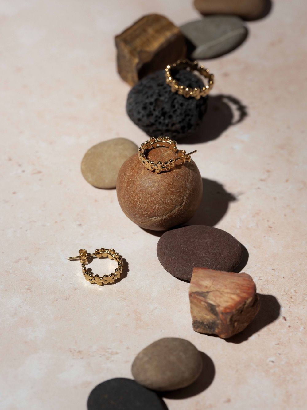 Natural river stones arranged with delicate gold jewelry pieces create an artistic composition on a textured beige surface.