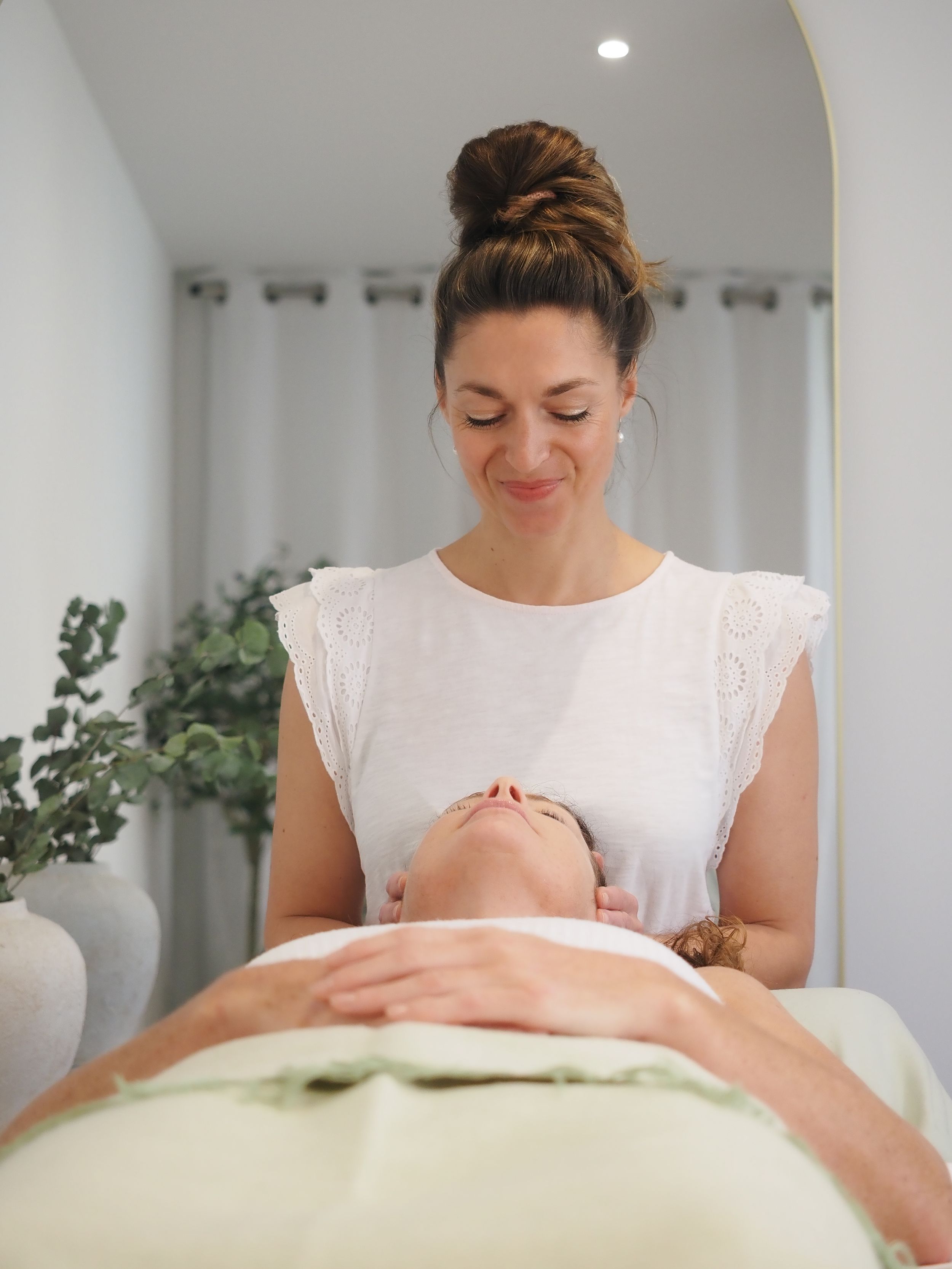 Massage therapist in white top providing relaxing facial treatment to client on massage table.