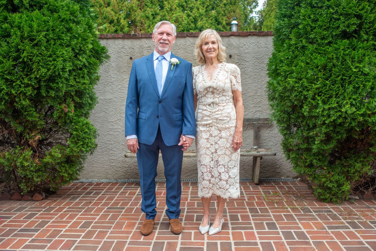 A couple stands on a brick walkway surrounded by green shrubbery in formal attire.