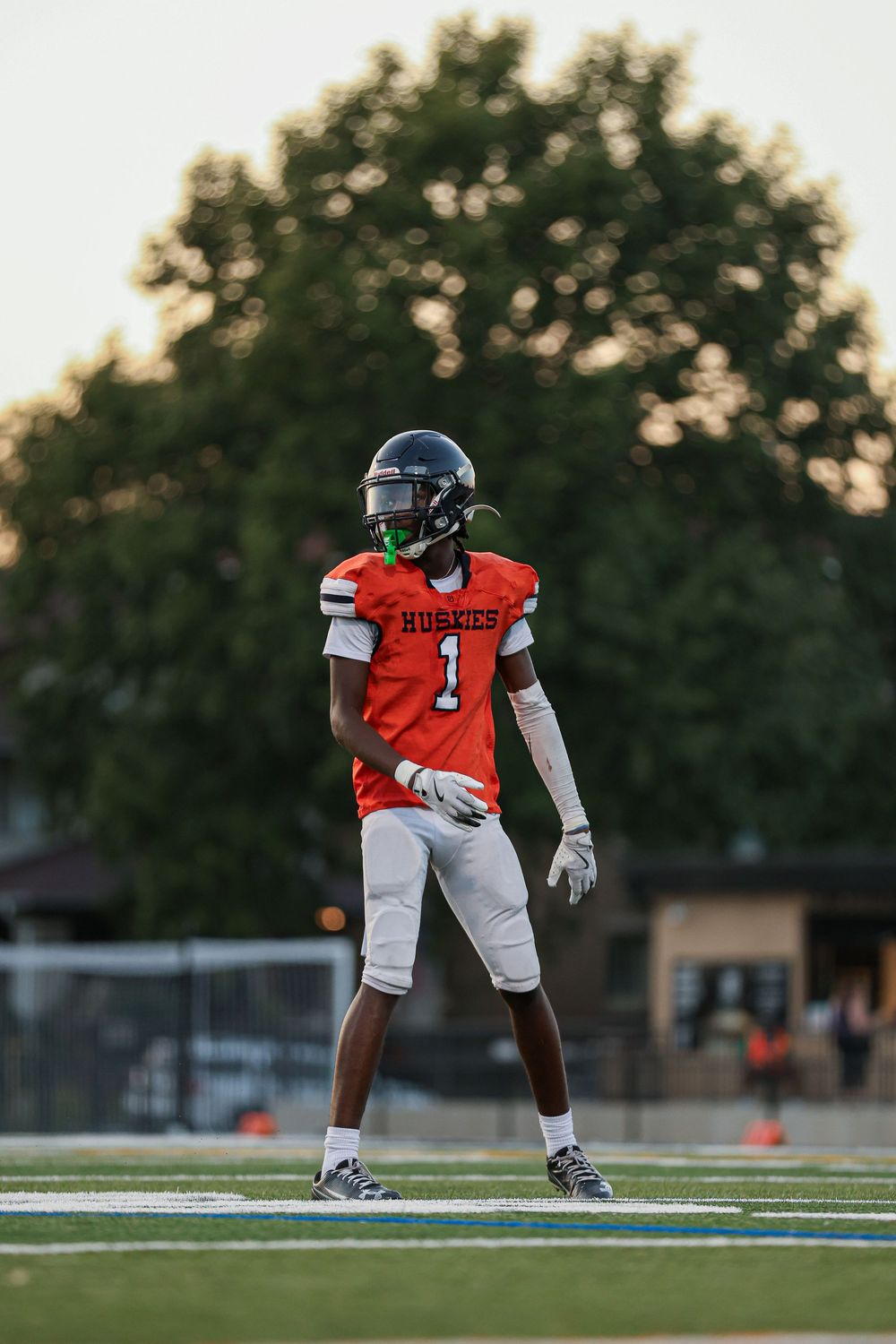Football player stands ready in orange jersey on field during evening practice session.