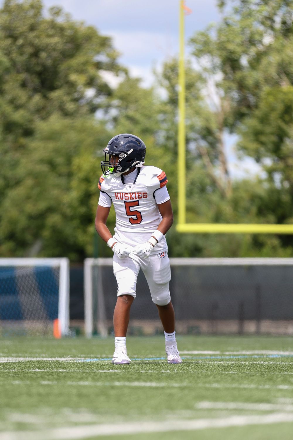 Football player in white uniform with number 5 stands ready on field with goal post visible in background.