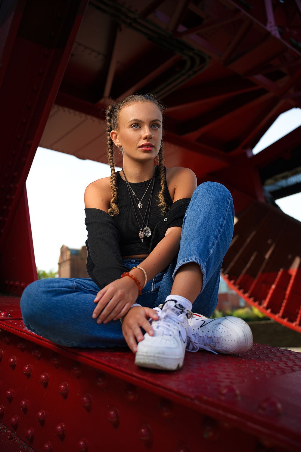 Person in black top and jeans sits on red surface wearing white sneakers and jewelry in moody lighting.