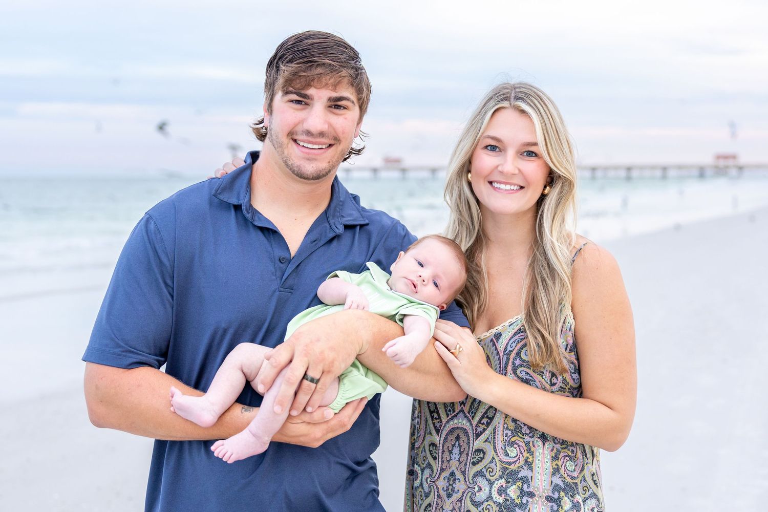 Young couple smiling on a beach at sunset while holding their newborn baby dressed in green.