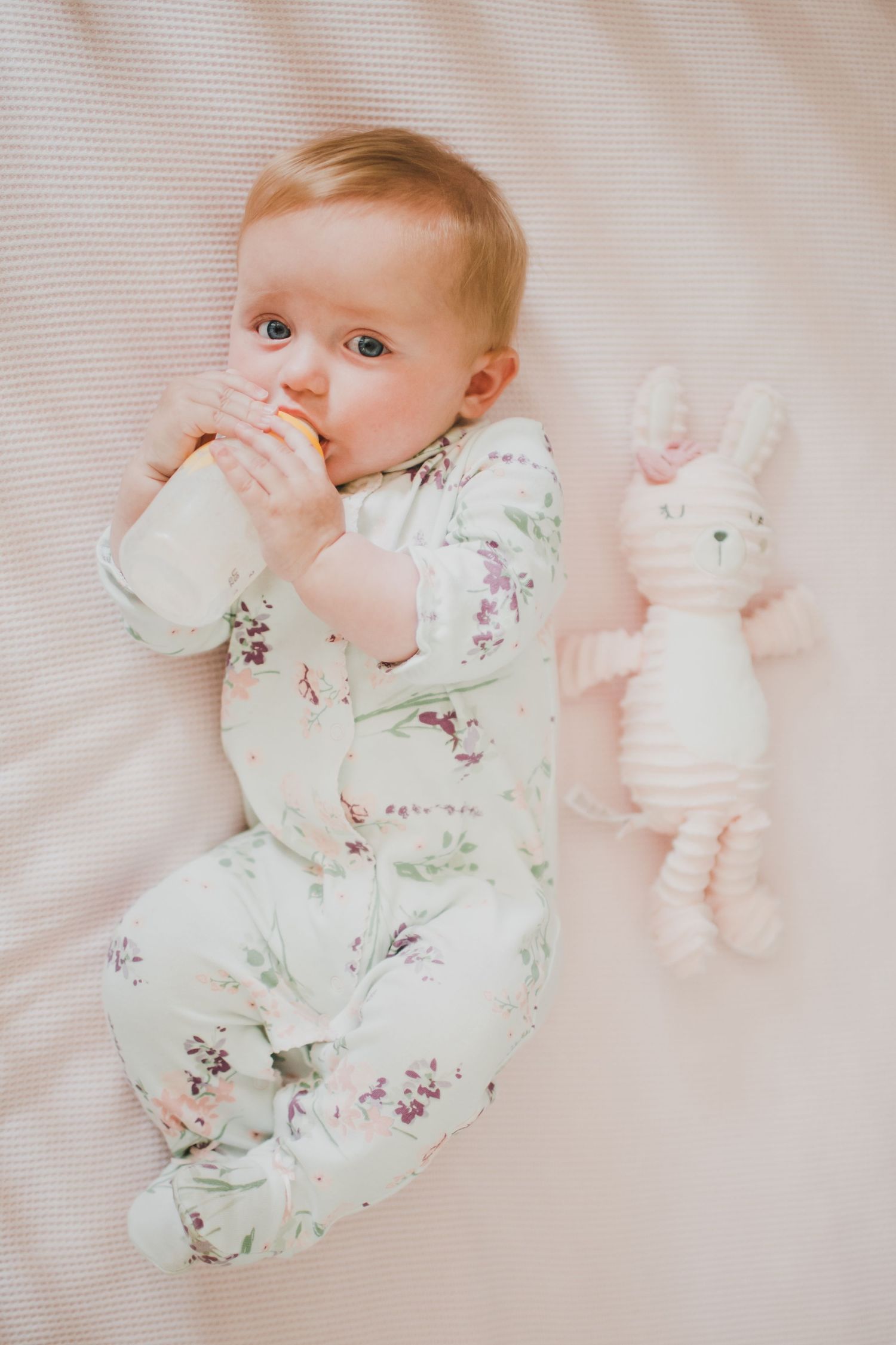 Baby in floral pajamas lying on white bedding with a stuffed bunny toy.