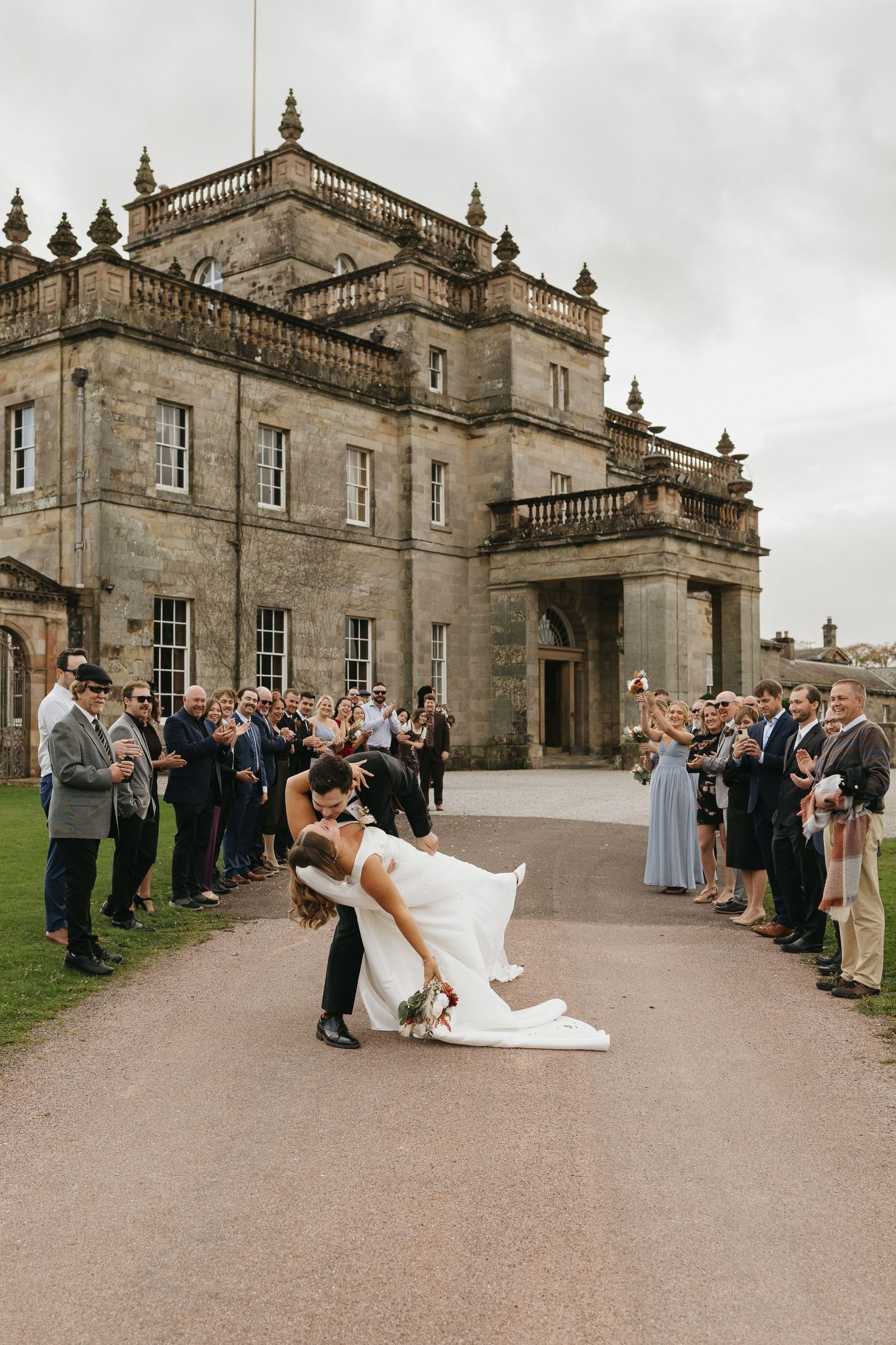 Wedding guests watch as newlyweds perform a romantic dip pose outside a grand historic stone mansion.