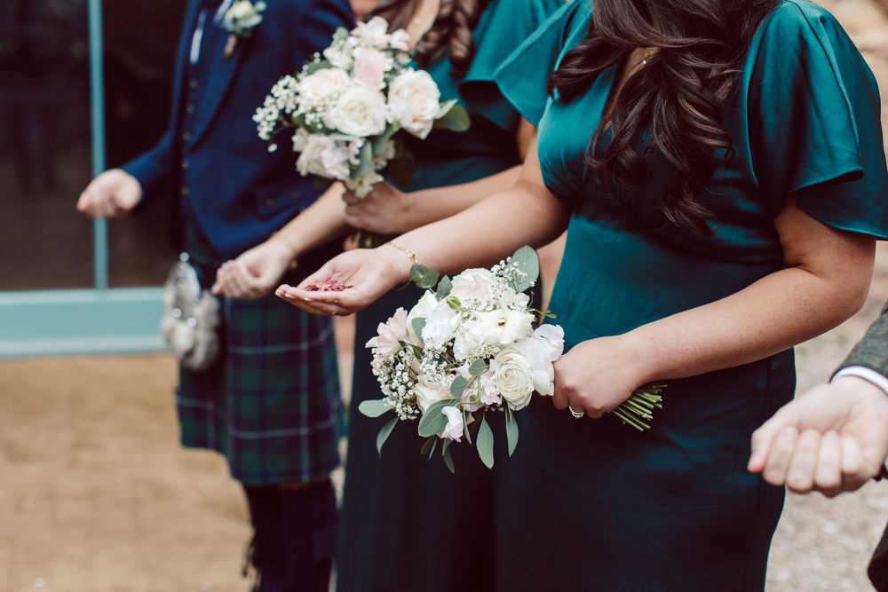 A couple holds hands during a wedding ceremony while exchanging rings, with white floral bouquets in view.