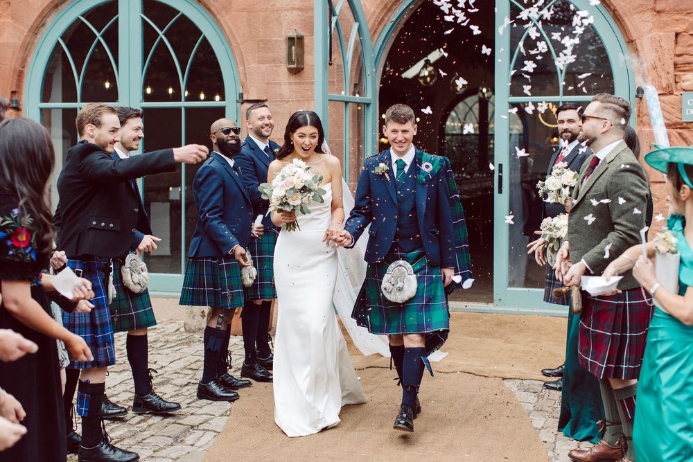 A wedding couple exits a Scottish church while guests in traditional kilts line the path throwing confetti.