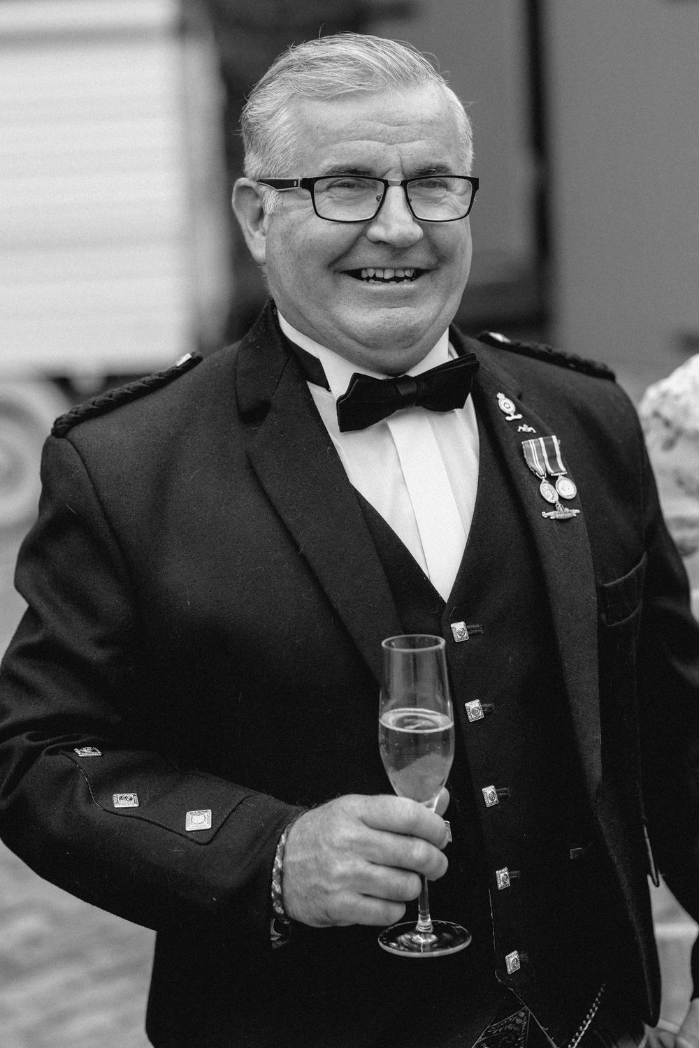 A person in formal black attire and glasses smiles while holding a champagne glass at an event.