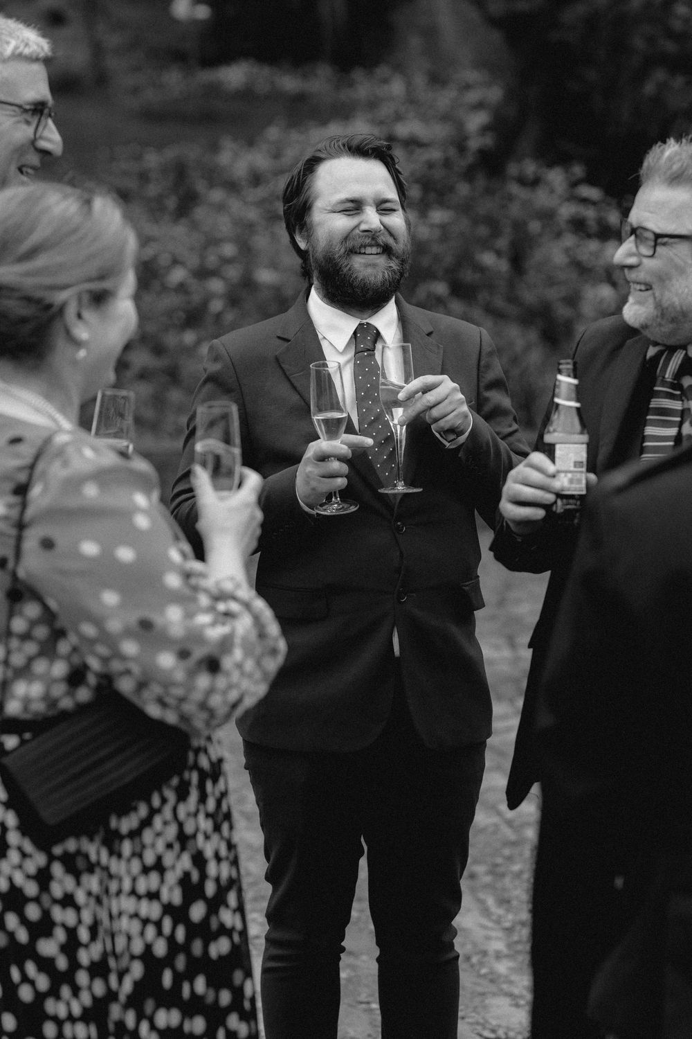 Wedding guests in formal attire share laughter while holding champagne glasses during an outdoor celebration.