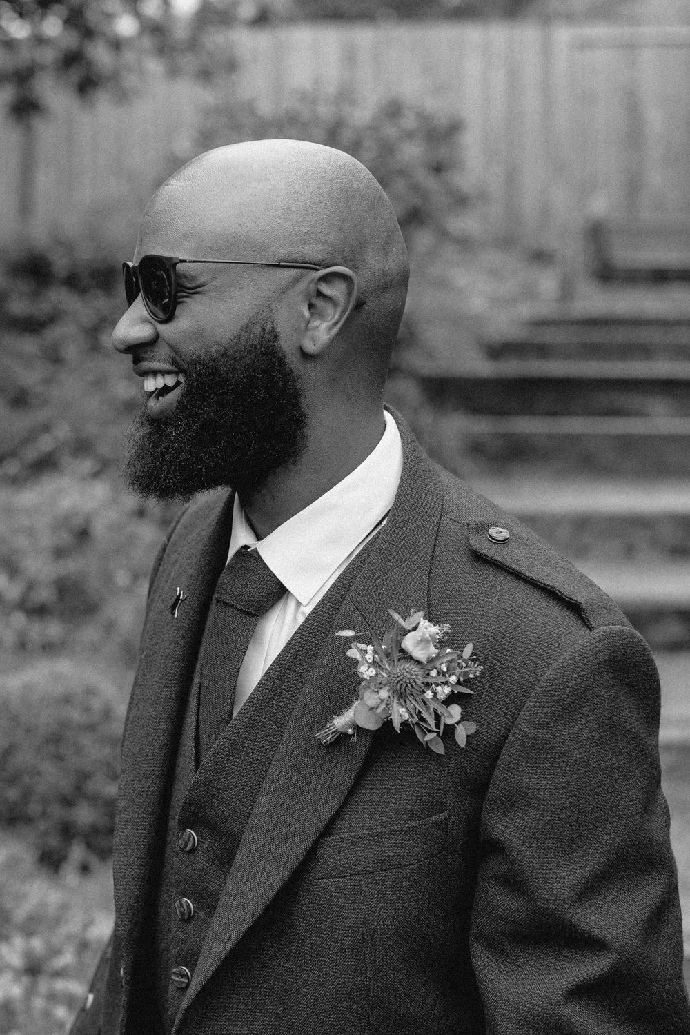Black and white portrait of a groom in a dark suit with boutonniere smiling during an outdoor wedding ceremony.