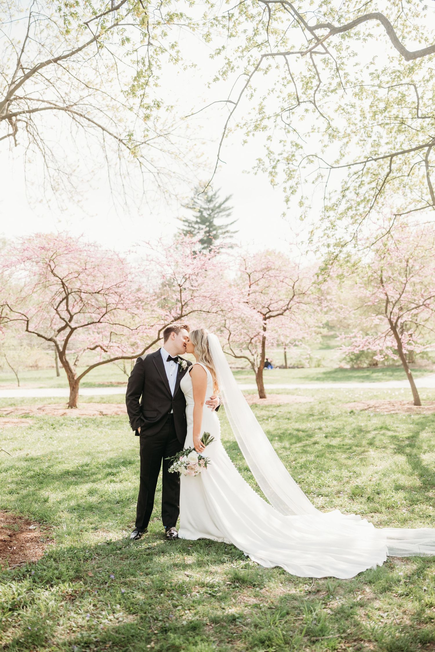 A couple in formal wedding attire walk through a scenic mountain meadow during autumn with sunlit trees in the background.