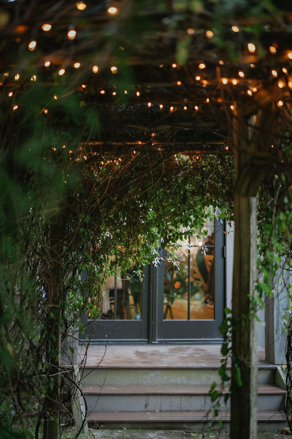 String lights illuminate a rustic outdoor patio covered in climbing vines and greenery at dusk.