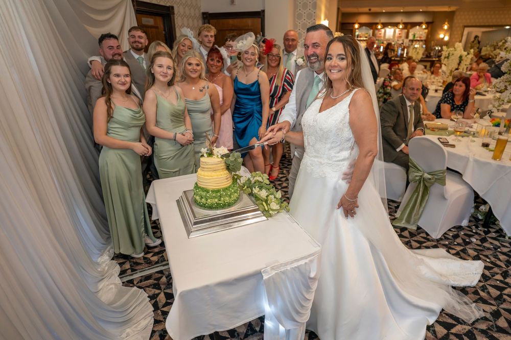 Wedding party gathered around a tiered cake with sage green bridesmaid dresses.