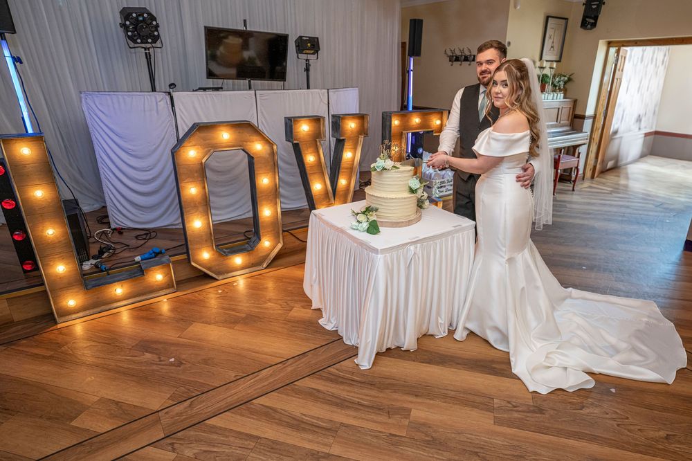 A wedding couple cuts their cake next to large illuminated LOVE letters at their reception venue.