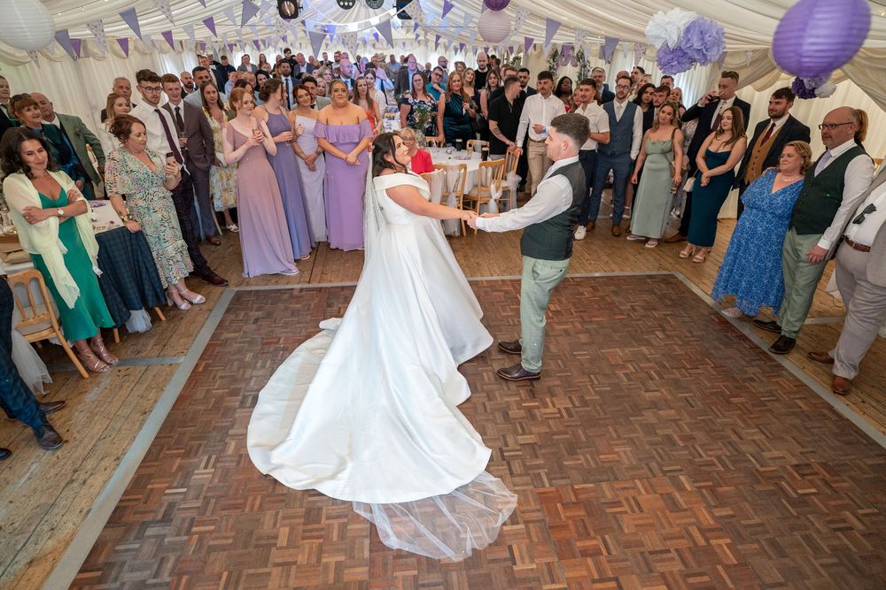 Wedding guests dance and celebrate on a wooden dance floor under a decorated white tent with purple lanterns.
