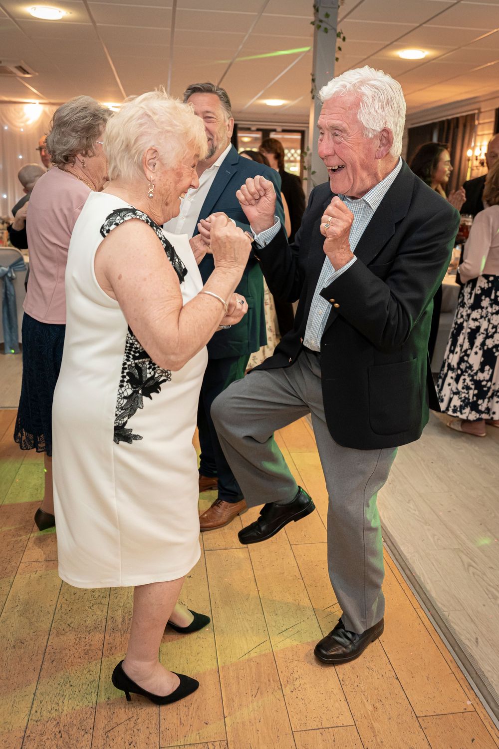 Elderly couples dancing and laughing together on a wooden dance floor at a celebration.
