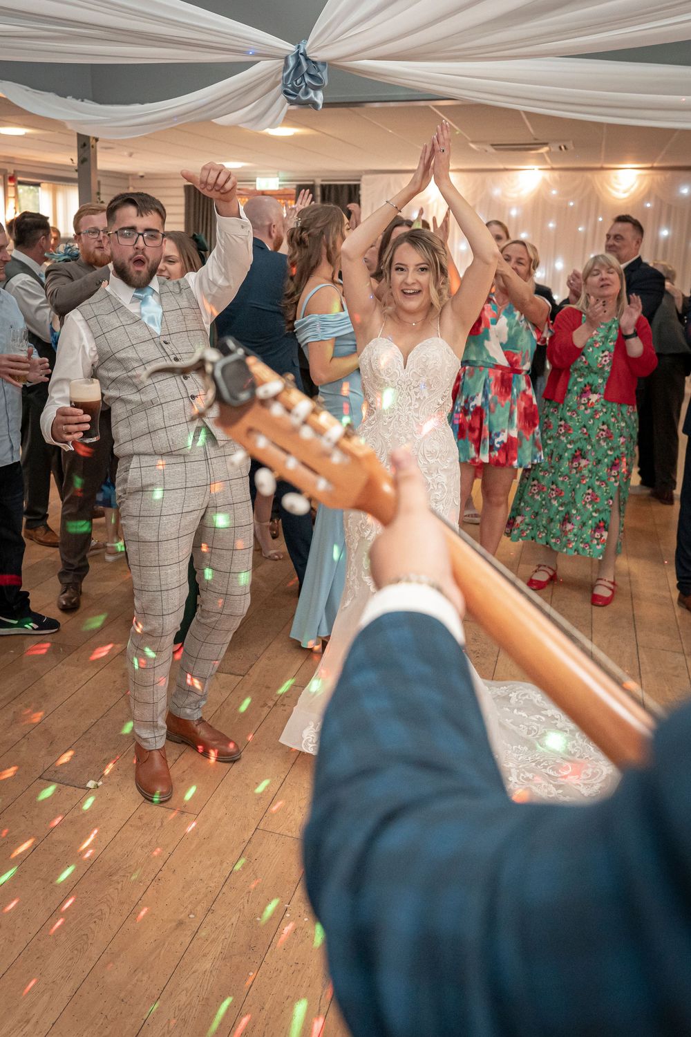 Wedding guests dancing energetically under draped fabric ceiling with colorful floor lighting.