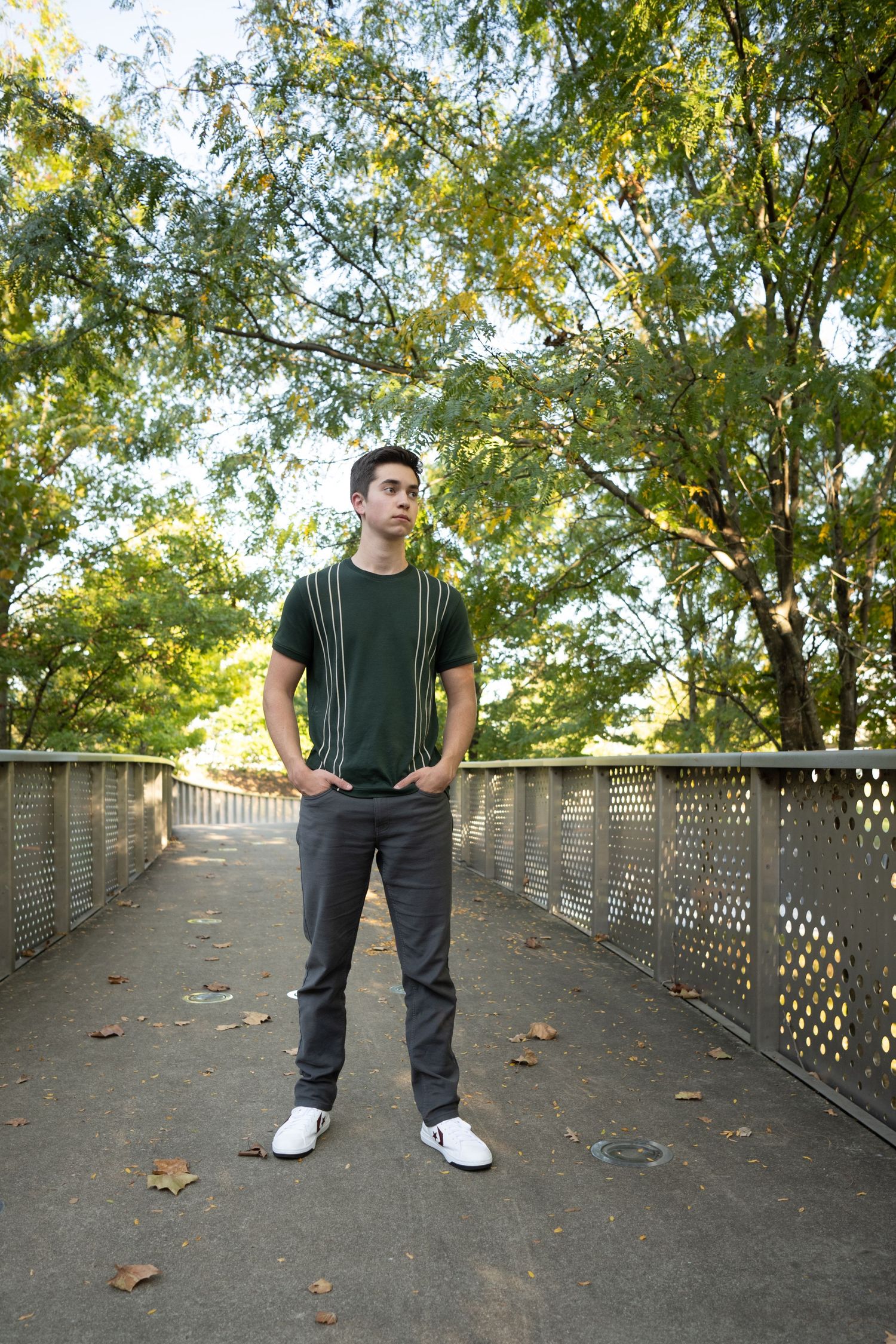 A person in casual attire stands on a wooden footbridge surrounded by autumn trees and fallen leaves.