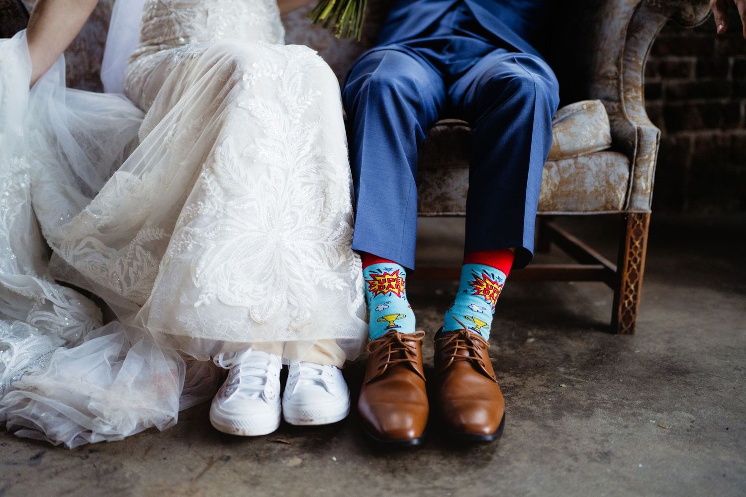 Bride and groom sitting showing off their feet, bride in white sneakers and groom in brown shoes with colorful fun socks.